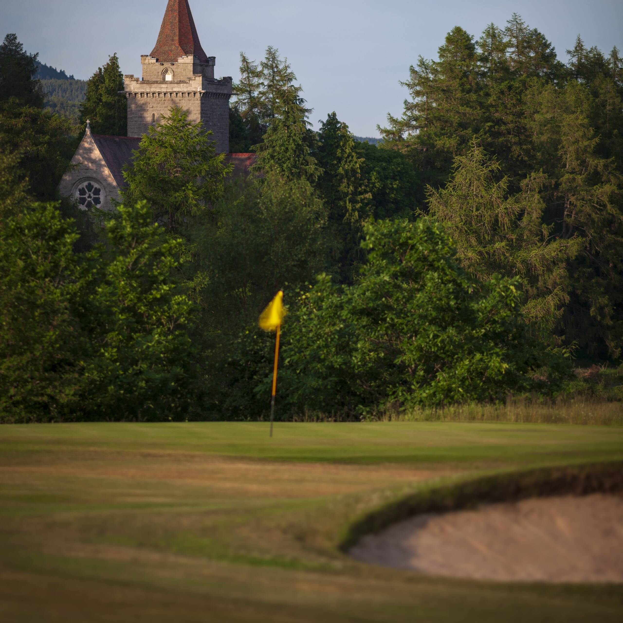 A view of the Balmoral Castle Golf Course with a yellow flag marking a hole in the foreground, surrounded by lush greenery and trees, with a stone tower visible in the background.